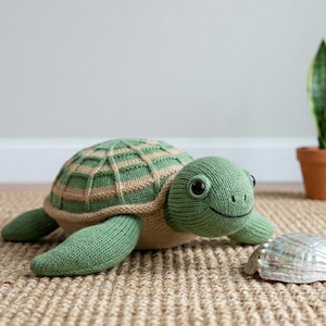 Pode incluir: Uma tartaruga de pelúcia de malha em tons de verde e bege, com um rosto sorridente e olhos grandes e pretos. A tartaruga está deitada em um tapete tecido, com uma concha por perto. Uma planta em vaso está ao fundo.