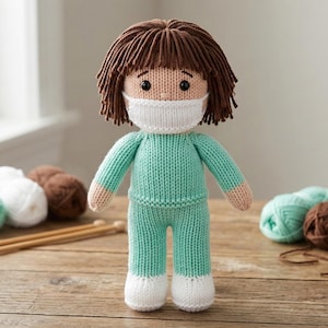 May include: A handmade knitted doll wearing a white face mask, teal scrubs, and white shoes. The doll has brown hair and is standing on a wooden surface. Knitting needles and yarn balls in white, teal, and brown are in the background.
