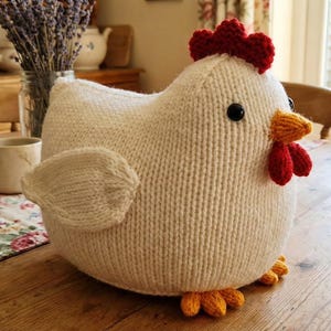 May include: A handmade, knitted chicken figurine. The plush chicken is primarily off-white with a red comb and wattle, black eyes, and orange beak and feet. The chicken is sitting on a wooden table.