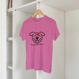 Puede incluir: Una camiseta rosa con un diseño de cara de perro de dibujos animados en negro. El perro tiene un corazón rojo con "ADHD" en la frente y el texto "My brain has zoomies" debajo. La camiseta está colgada en una percha de madera.