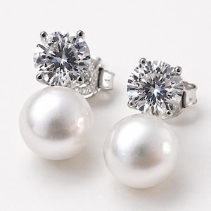 May include: A pair of stud earrings. Each earring has a large, clear, round-cut stone above a white, round pearl. The earrings have a silver-coloured metal setting and a post-and-clutch closure.