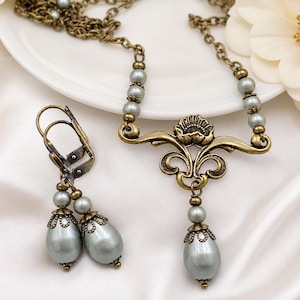 Pode incluir: Um conjunto de joias composto por um colar e brincos a condizer. O colar apresenta uma corrente em tom de bronze com contas de p&eacute;rolas cinzentas e um pendente floral central. Os brincos s&atilde;o pendentes com fecho de alavanca.