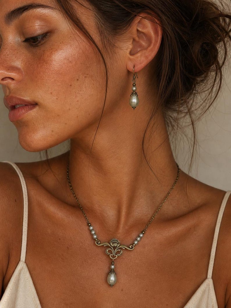 Pode incluir: Um conjunto de joias composto por um colar e um brinco. O colar apresenta uma corrente em tom de bronze com pequenas p&eacute;rolas cinzentas e um pendente central de bronze ornamentado com uma p&eacute;rola cinzenta pendente a combinar.