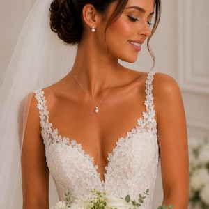 May include: A white lace wedding dress featuring a deep V-neckline with intricate floral embroidery, thin straps, and a sheer tulle veil. The ensemble is accessorized with a delicate silver chain necklace with a single pearl pendant and matching pearl earrings.