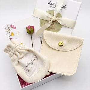Pode incluir: Uma caixa de presente branca contendo uma bolsa de veludo creme com cord&atilde;o e a marca A&U Jewelry, uma bolsa de veludo creme a condizer com bot&atilde;o de press&atilde;o, uma pequena rosa seca cor-de-rosa e um cart&atilde;o branco com flores.