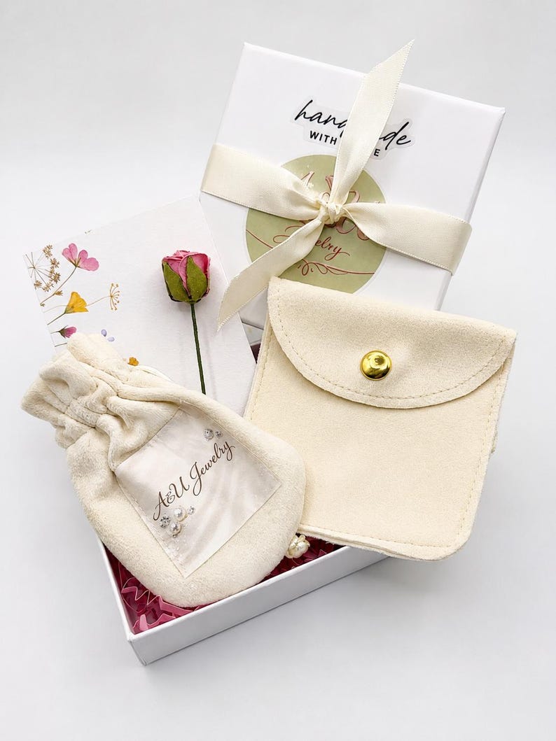 Pode incluir: Uma caixa de presente branca contendo uma bolsa de veludo creme com cord&atilde;o e a marca A&U Jewelry, uma bolsa de veludo creme a condizer com bot&atilde;o de press&atilde;o, uma pequena rosa seca cor-de-rosa e um cart&atilde;o branco com flores.