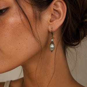 Pode incluir: Um brinco pendente com uma conta lisa em forma de gota na cor verde s&aacute;lvia, suspensa por um gancho de metal em tom de bronze. A conta &eacute; acentuada com uma pequena conta redonda a condizer e uma tampa de metal decorativa.