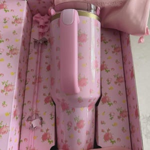 May include: A pink floral Stanley tumbler with a handle, presented in a matching box. The tumbler features a pink and gold rim, a pink handle, and a floral pattern. The box and accessories, including a pink bag and stir sticks, complement the tumbler's design.