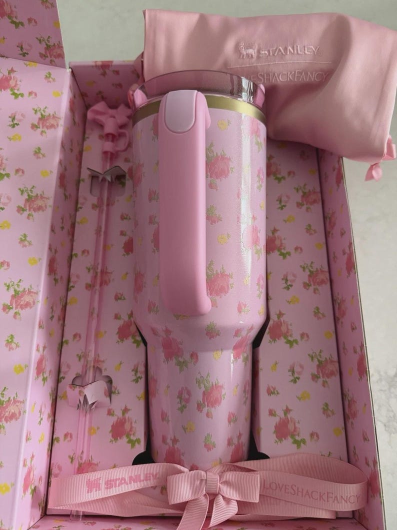 May include: A pink floral Stanley tumbler with a handle, presented in a matching box. The tumbler features a pink and gold rim, a pink handle, and a floral pattern. The box and accessories, including a pink bag and stir sticks, complement the tumbler's design.