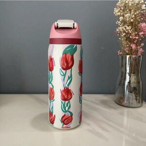 Puede incluir: Una botella de agua aislada blanca con tapa rosa y un acento granate. La botella est&aacute; decorada con un patr&oacute;n repetido de tulipanes rojos con hojas verdes y acentos morados y azules. Tiene una tapa abatible.