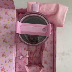 May include: A pink Stanley tumbler with a matching lid and strap, presented in a floral-patterned box. The box and accessories are in shades of pink, with the Stanley logo visible on the lid and a matching cloth.