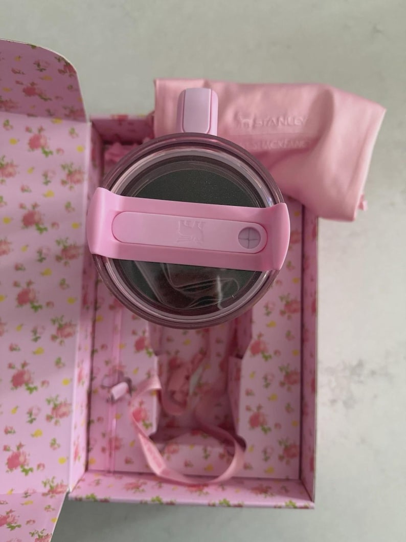 May include: A pink Stanley tumbler with a matching lid and strap, presented in a floral-patterned box. The box and accessories are in shades of pink, with the Stanley logo visible on the lid and a matching cloth.