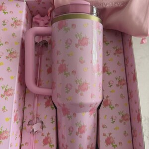 May include: A pink floral Stanley tumbler with a handle, presented in a matching floral box. The tumbler has a pink lid and gold accents. A pink bow and a pink bag with the Stanley logo are also visible.