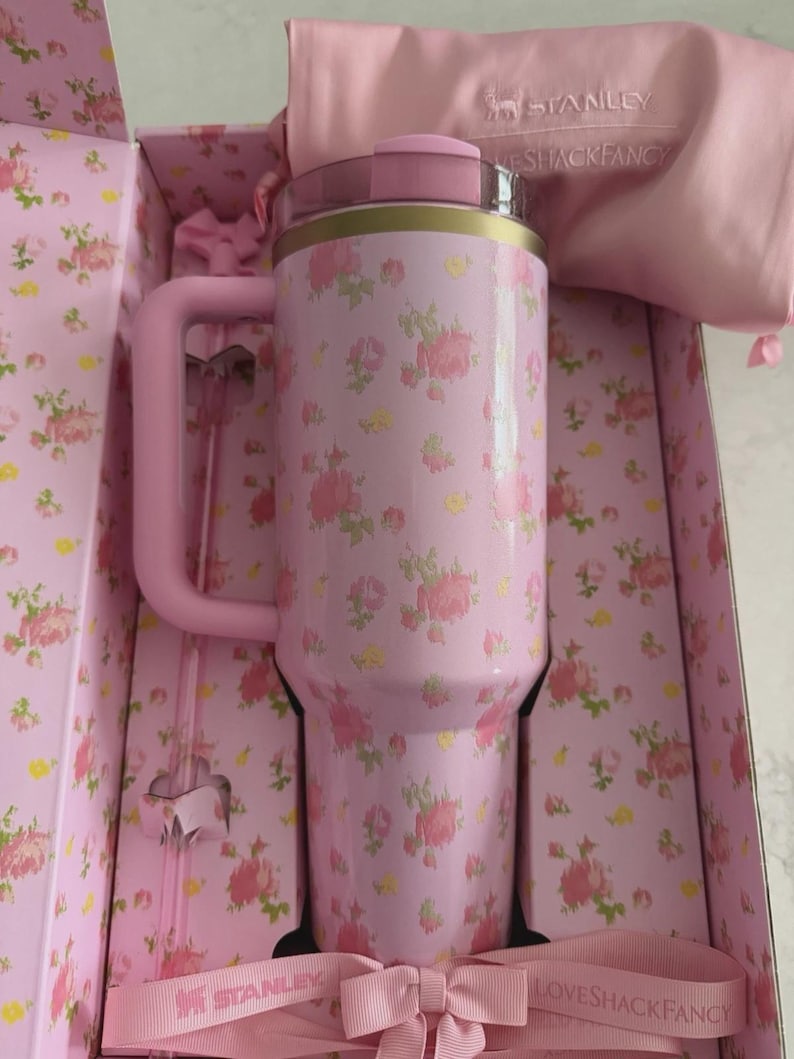 May include: A pink floral Stanley tumbler with a handle, presented in a matching floral box. The tumbler has a pink lid and gold accents. A pink bow and a pink bag with the Stanley logo are also visible.