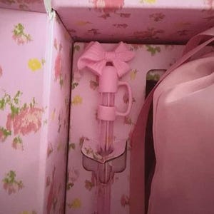May include: A pink bottle with a bow-shaped stopper is inside a floral-patterned box. The box and bottle are a soft pink color. The box has a floral pattern with pink and yellow flowers.
