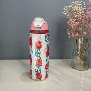 Puede incluir: Una botella de agua estampada con tapa rosa y burdeos. La botella presenta un fondo blanco con ilustraciones de tulipanes rojos y hojas verdes. La tapa tiene el texto "Lift to Unlock".