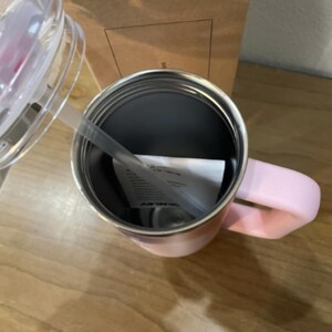 以下が含まれることがあります： ステンレス製の内部、同色のピンクのハンドル、ストロー付きの透明なプラスチック製の蓋を備えたライトピンクの断熱タンブラー。カップの内側には黒い文字が書かれた小さな白い紙のインサートが見え、縁は金属製です。