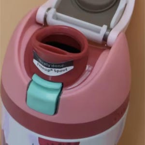 Puede incluir: Primer plano de una botella de agua rosa y blanca con un dise&ntilde;o floral. La botella tiene una tapa abatible con una boquilla gris y un pestillo turquesa. El texto "Nicely done. FreeSip Spout" est&aacute; impreso en la boquilla.