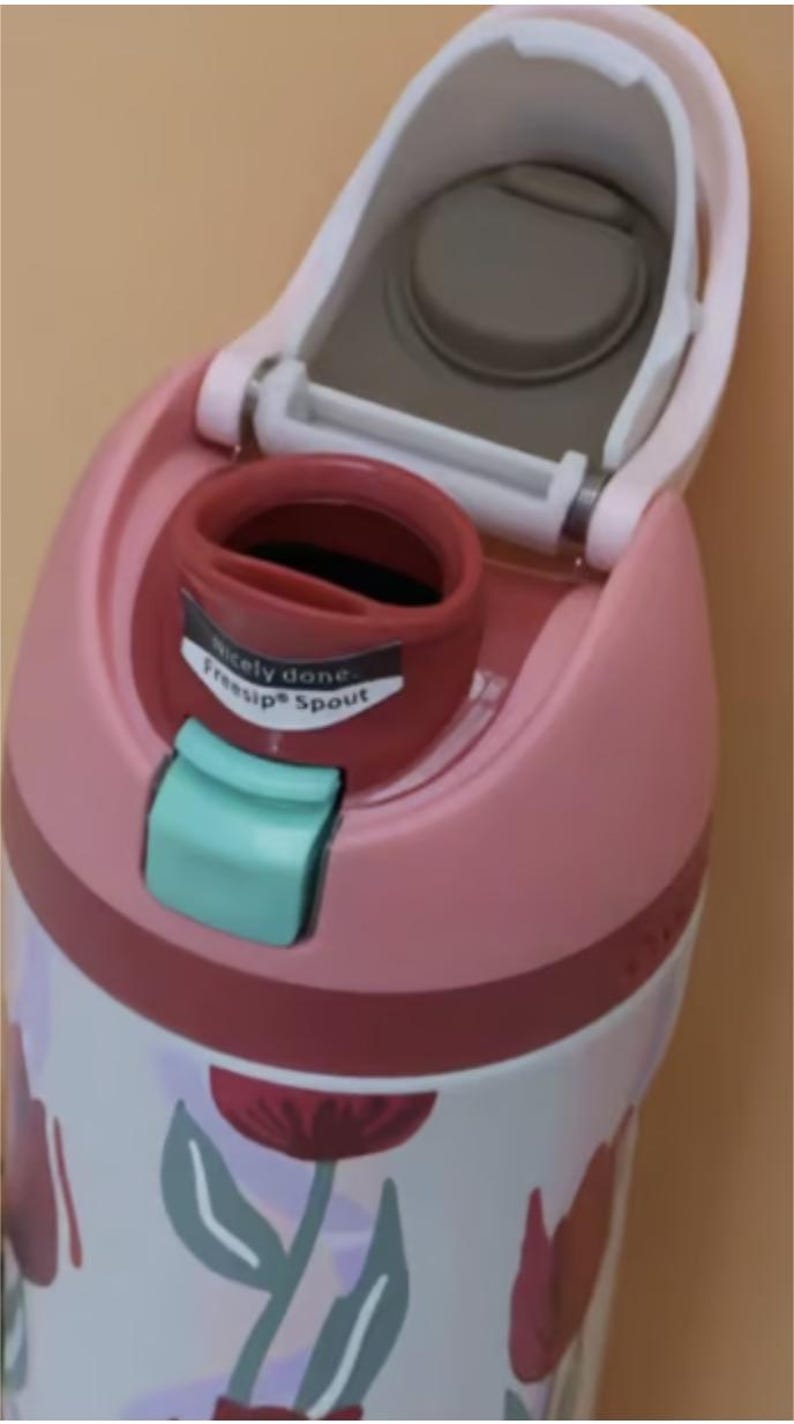 Puede incluir: Primer plano de una botella de agua rosa y blanca con un dise&ntilde;o floral. La botella tiene una tapa abatible con una boquilla gris y un pestillo turquesa. El texto "Nicely done. FreeSip Spout" est&aacute; impreso en la boquilla.