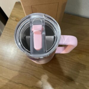 以下が含まれることがあります： 淡いピンク色のステンレス製タンブラー。スライド式の開閉機構を備えた透明なプラスチック製の蓋と、同色のピンクのハンドルが付いています。タンブラーは直立しており、蓋の上面と側面のハンドル部分が見えています。