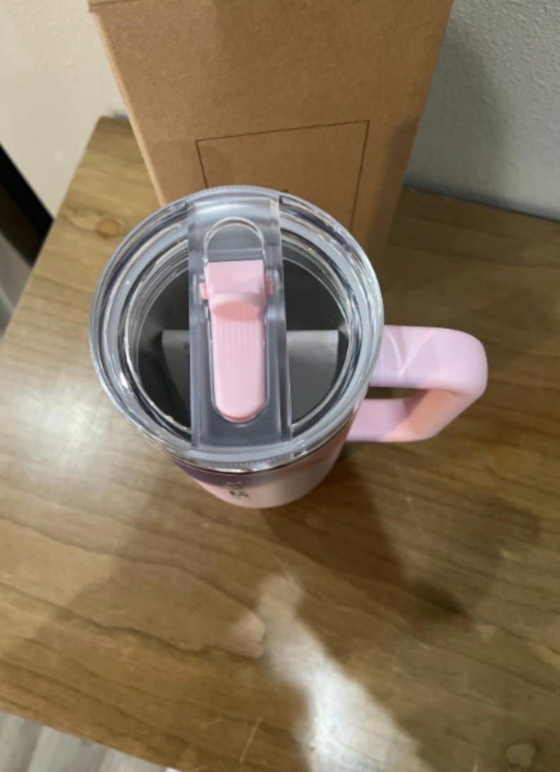 以下が含まれることがあります： 淡いピンク色のステンレス製タンブラー。スライド式の開閉機構を備えた透明なプラスチック製の蓋と、同色のピンクのハンドルが付いています。タンブラーは直立しており、蓋の上面と側面のハンドル部分が見えています。
