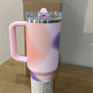 以下が含まれることがあります： ハンドル付きの背の高いステンレス製断熱タンブラー。ソフトピンク、ピーチ、パープルの抽象的なグラデーション仕上げが特徴です。透明なプラスチック製の蓋にはピンクのスライド式開閉口があり、底面には白い製品ラベルが巻き付けられています。