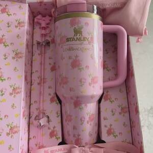 May include: A pink Stanley tumbler with a floral pattern, presented in a matching box. The tumbler has a handle and a clear lid. The box and accessories, including a straw and ribbon, complement the tumbler's design. The text "STANLEY" and "LOVESHACKFANCY" are visible.