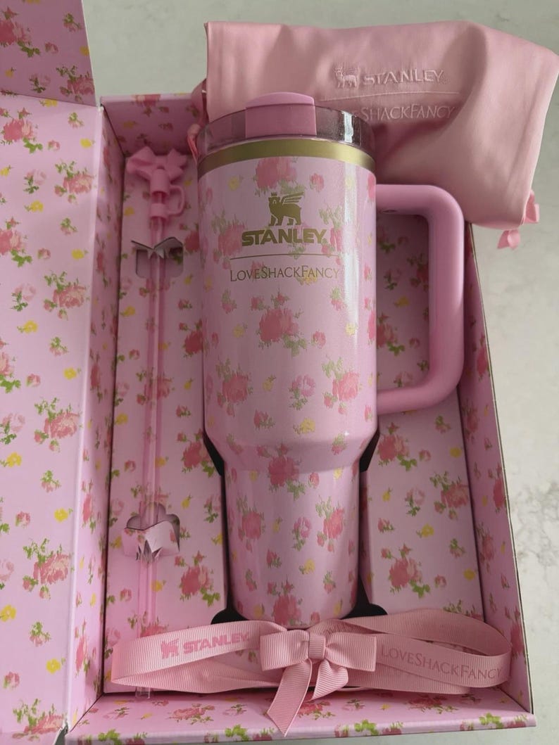 May include: A pink Stanley tumbler with a floral pattern, presented in a matching box. The tumbler has a handle and a clear lid. The box and accessories, including a straw and ribbon, complement the tumbler's design. The text "STANLEY" and "LOVESHACKFANCY" are visible.