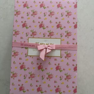 May include: A rectangular pink box with a floral pattern of pink roses and green leaves. The box is wrapped with a pink ribbon and bow. The text "STANLEY" and "LOVESHACKFANCY" are visible on the ribbon.