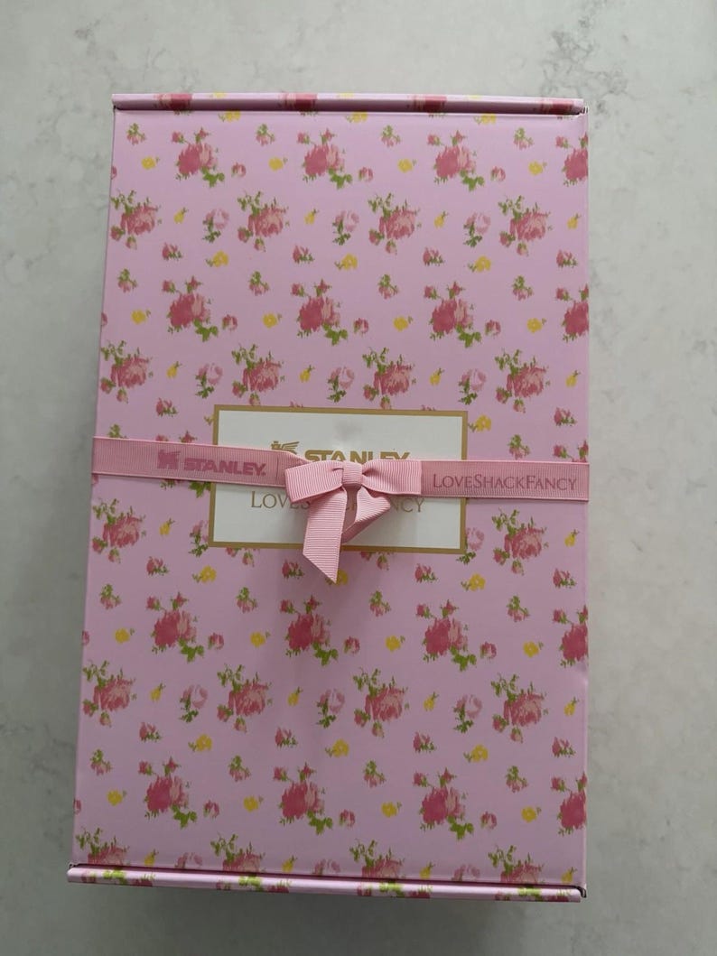May include: A rectangular pink box with a floral pattern of pink roses and green leaves. The box is wrapped with a pink ribbon and bow. The text "STANLEY" and "LOVESHACKFANCY" are visible on the ribbon.