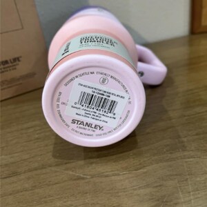 以下が含まれることがあります： ハンドル付きのライトピンクのスタンレー クエンチャー H2.0 FlowState タンブラー。底面にはバーコード付きの白い製品ラベルがあり、Stanley A Brand of PMIの文字と、40オンス、1.18リットル、BPAフリー、食器洗浄機対応という情報が記載されています。