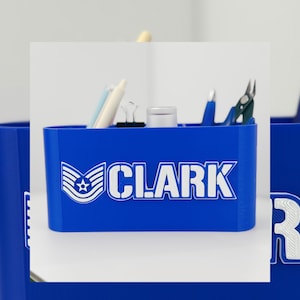 Peut inclure: Un organiseur de bureau bleu avec le nom "CLARK" en lettres blanches et un emblème militaire. L'organisateur contient des stylos, une pince à reliure et d'autres petites fournitures de bureau. L'organisateur est en plastique.