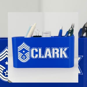 Może przedstawiać: Niebieski organizer biurkowy z białym napisem "CLARK". Organizer posiada emblemat wojskowy i mieści długopisy, mały pojemnik i inne artykuły biurowe. Tło jest białe.