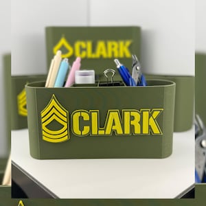 Puede incluir: Organizador de escritorio verde oliva con texto amarillo e insignia militar. El organizador está lleno de bolígrafos, lápices y otros útiles de oficina. El texto en el organizador dice "CLARK".