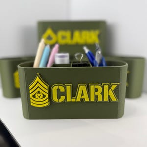 Puede incluir: Organizador de escritorio verde oliva con el nombre "CLARK" en letras amarillas. El organizador tiene múltiples compartimentos y contiene bolígrafos, lápices y otros útiles de oficina. También se muestra una insignia militar en amarillo.