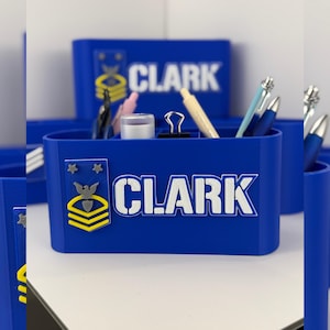 May include: A blue desk organizer with the name "CLARK" in white lettering. The organizer has a military emblem and holds pens, pencils, and other office supplies. The design is 3D printed.