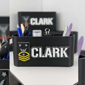 Può includere: Organizzatore da scrivania nero con il nome "CLARK" a lettere bianche. Presenta un emblema in stile militare con stelle e un design a galloni giallo e oro. All'interno sono presenti penne, matite e altri articoli per l'ufficio.