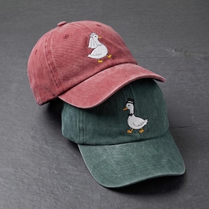 Op de afbeelding: Twee baseballpetten in rood- en groentinten. Elke pet heeft een geborduurd ganzenontwerp. De rode pet heeft een gans met een bruidssluier, de groene pet een gans met een hoge hoed.