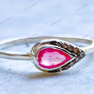 May include: A silver ring featuring a central pear-shaped pink gemstone surrounded by a halo of small, dark brown accent stones. The band is smooth and metallic, showcasing a teardrop-shaped setting that holds the vibrant pink center stone securely.