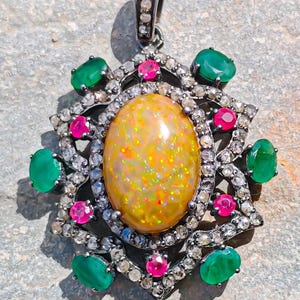 May include: A pendant featuring a large oval yellow opal centre stone surrounded by a decorative frame of small clear stones, four oval green stones, and four round pink stones. The metal setting is dark, and a bail is attached at the top.