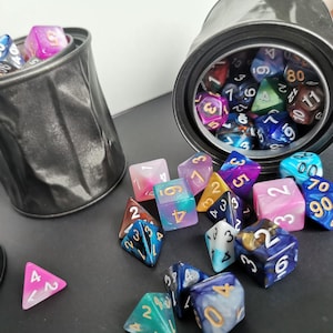 May include: A collection of colorful polyhedral dice, including D4, D6, D8, D10, D12, and D20, are scattered around two dented, black metal containers. The dice feature various colors and patterns, with visible numbers.