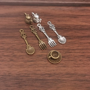 Puede incluir: Colección de abalorios metálicos en miniatura, que incluyen una taza y platillo, cucharas, tenedores y dos pequeñas figuras. Los abalorios son en tonos oro y plata antiguos, dispuestos sobre una superficie de madera oscura.