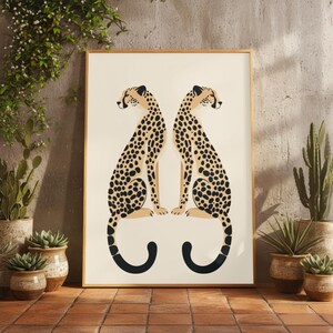 May include: Framed art print featuring two stylized cheetahs facing each other. The cheetahs have tan bodies with black spots and black tails. The background is off-white, and the frame is a light brown color. The print is displayed on a brick surface.