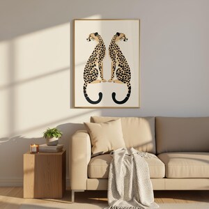 May include: A framed art print featuring two stylized cheetahs facing each other. The cheetahs have black spots on a tan body, with black curved tails. The artwork is in a gold frame, displayed above a beige sofa.