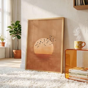 Pode incluir: Uma impressão de arte de parede retangular emoldurada com um grande sol laranja pálido centrado num fundo quente de cor terracota. Um bando de pequenos pássaros pretos é retratado a voar sobre o sol e a área do céu circundante.