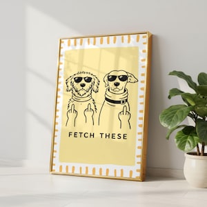 Könnte beinhalten: Gerahmter Kunstdruck mit gelbem Hintergrund, der zwei Zeichentrickhunde mit Sonnenbrillen zeigt, die den Mittelfinger zeigen. Der Text "FETCH THESE" steht darunter, und der Rahmen ist goldfarben.