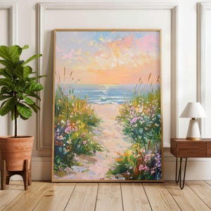 May include: An oil painting in a wooden frame shows a beach path leading to the sea at sunset. The path is bordered by wildflowers and grasses. The artwork is displayed on a light wood floor, with a potted plant and lamp nearby.
