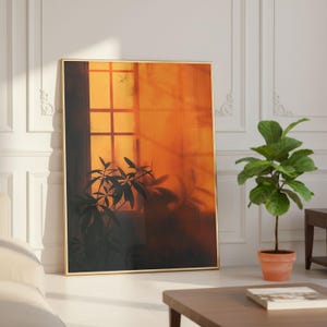 May include: A framed art print with a gold frame, depicting an orange-hued window scene with plant silhouettes. The artwork leans against a white wall. A potted plant and a coffee table are in the foreground, creating a warm, inviting atmosphere.