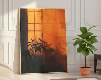 May include: A framed art print featuring a warm, orange-toned window scene with silhouetted plants. The artwork has a gold frame and is leaning against a white wall. A potted plant and a coffee table are in the foreground.