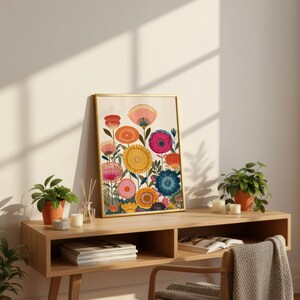 May include: A framed floral art print featuring colorful stylized flowers in shades of pink, yellow, orange, and blue. The artwork is displayed on a light wood console table, with small potted plants and candles adding to the decor. The frame has a gold finish.